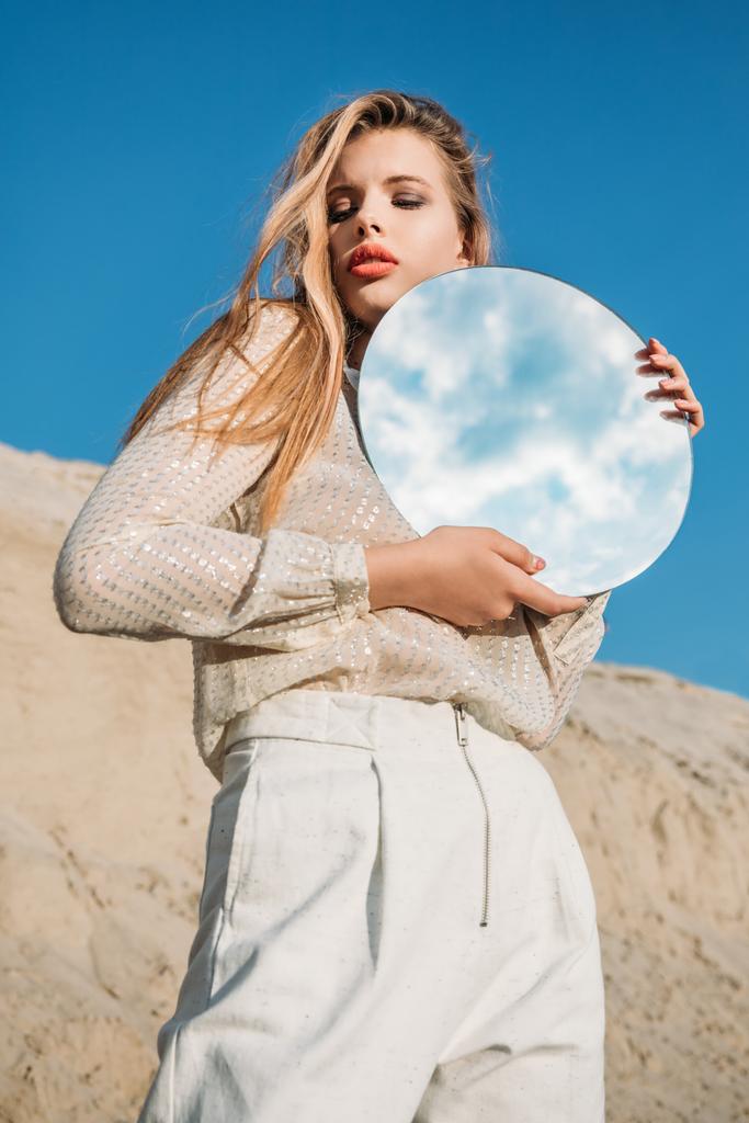 modello biondo alla moda in vestiti bianchi che tengono lo specchio rotondo con riflessione di cielo nuvoloso
 - Foto, immagini