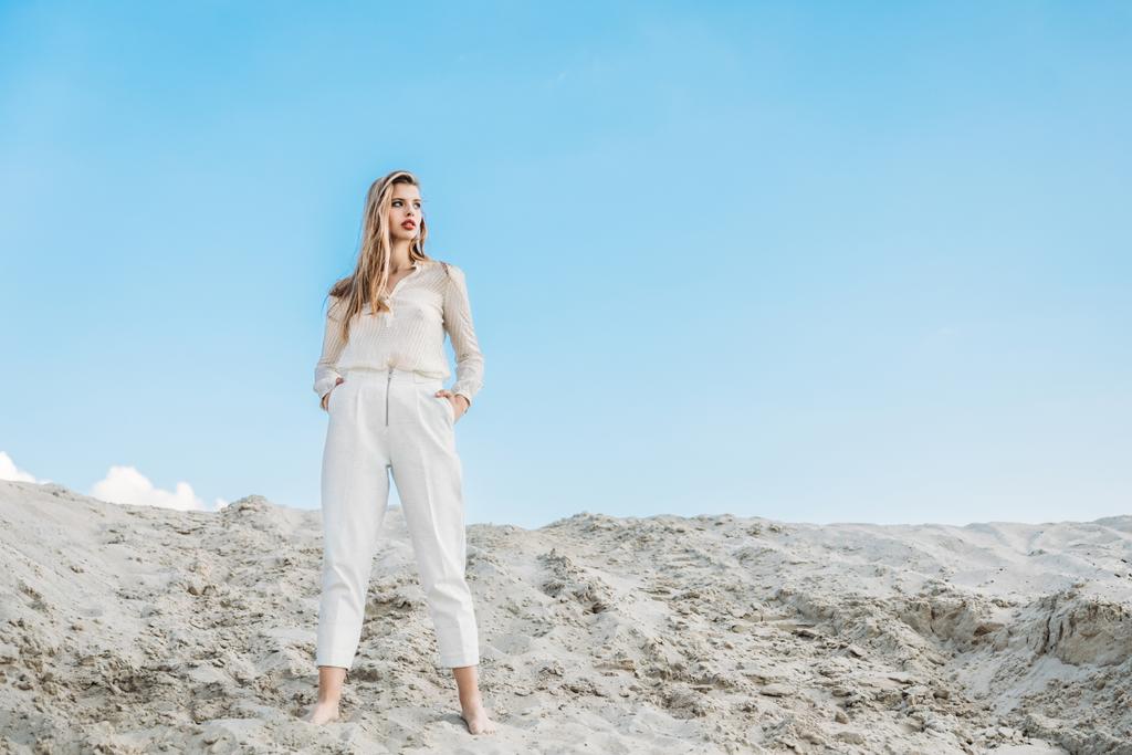 κομψή γυναίκα με λευκά ρούχα, ποζάρει στον αμμόλοφο με το μπλε του ουρανού - Φωτογραφία, εικόνα