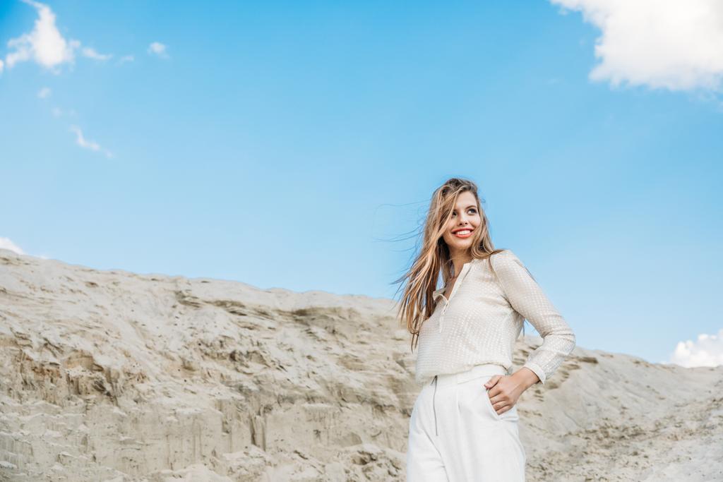houkutteleva hymyilevä nainen valkoisissa tyylikkäissä vaatteissa poseeraa hiekkadyynillä sinisellä taivaalla
 - Valokuva, kuva