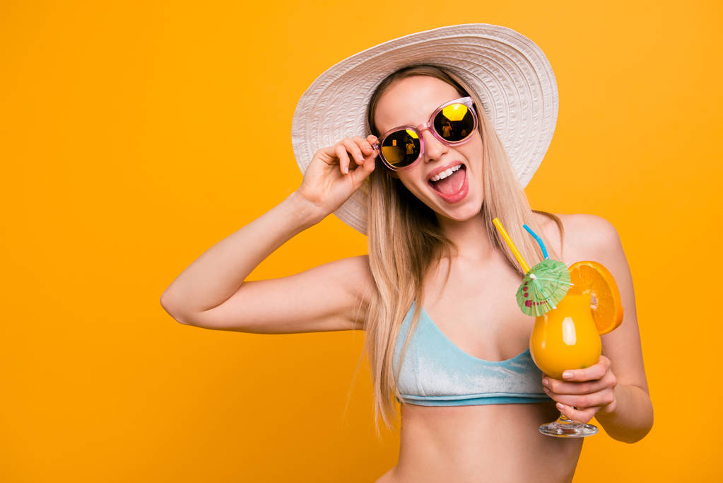Приваблива мила пряме волосся красива блондинка біла кавказька дівчина, одягнена в синій купальник, сонцезахисні окуляри, капелюх. Відкритий рот, схвильований, тримає склянку апельсинового коктейлю. Ізольовані на жовтому тлі
 - Фото, зображення