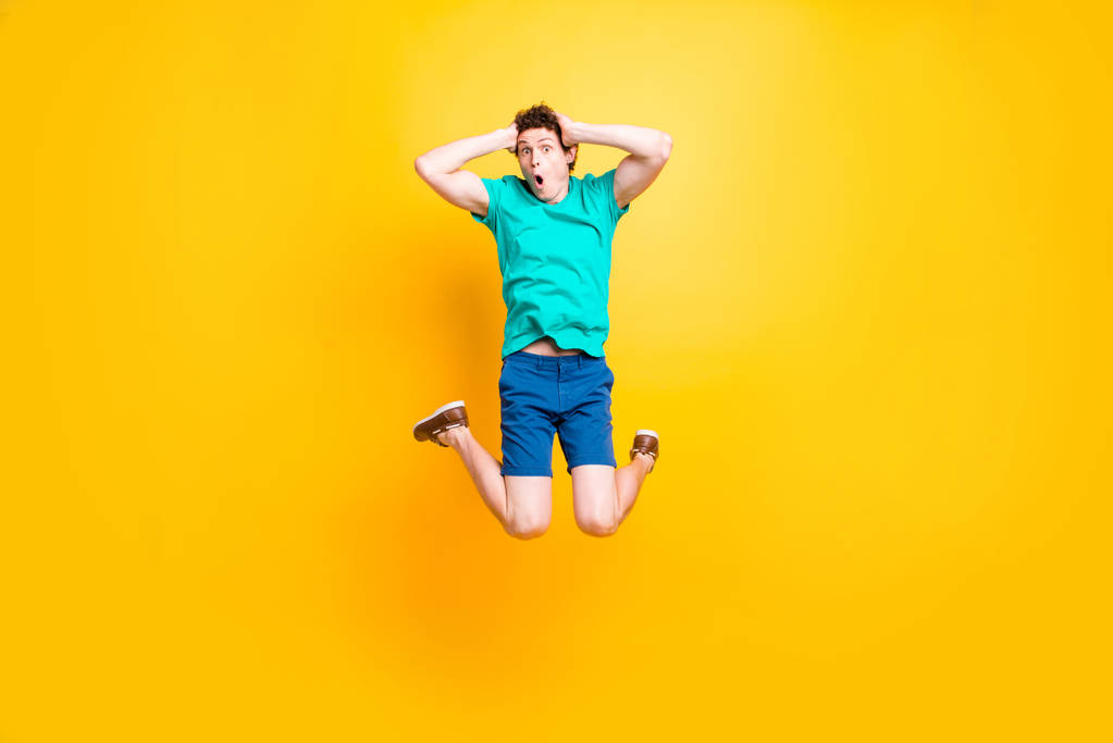Young attractive handsome funky curly-haired excited guy wearing casual green polo, shorts, sneakers, jumping up in the air, holding hands on head. Isolated over vivid shine bright yellow background - Photo, Image