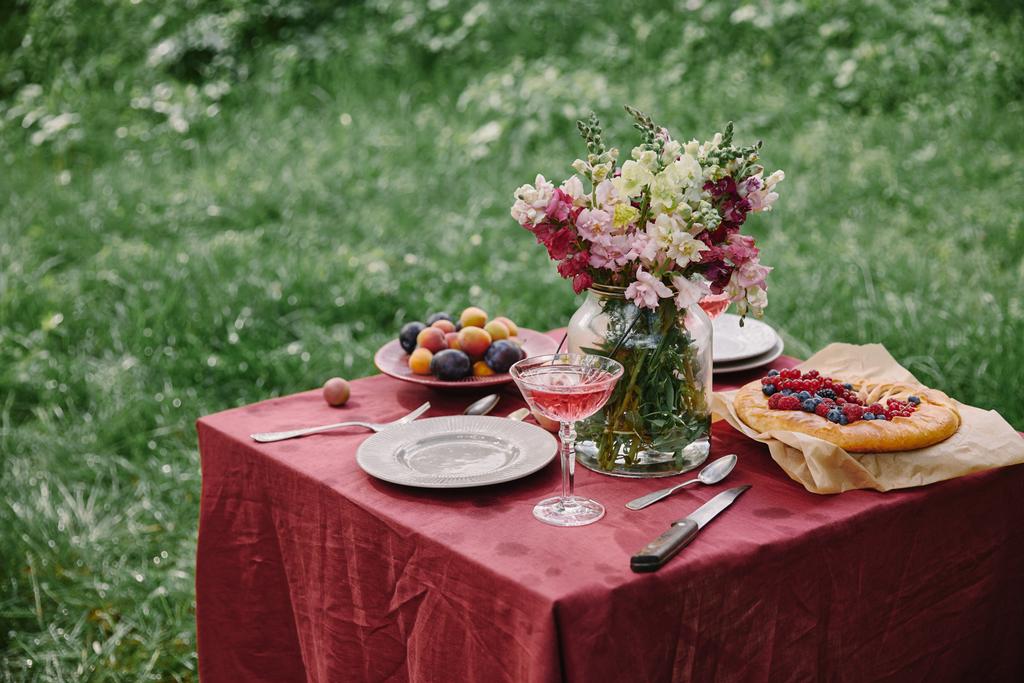 wineglass, berries pie and bouquet of flowers on table in green garden  - Photo, Image