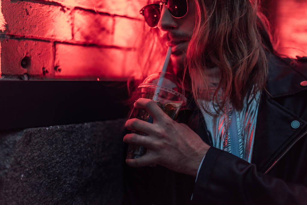 ελκυστικό νεαρό άνδρα σε γυαλιά ηλίου και δερμάτινο σακάκι πίνοντας Πάρτε μακριά κοκτέιλ από πλαστικό φλιτζάνι κάτω από το κόκκινο φως στο δρόμο - Φωτογραφία, εικόνα