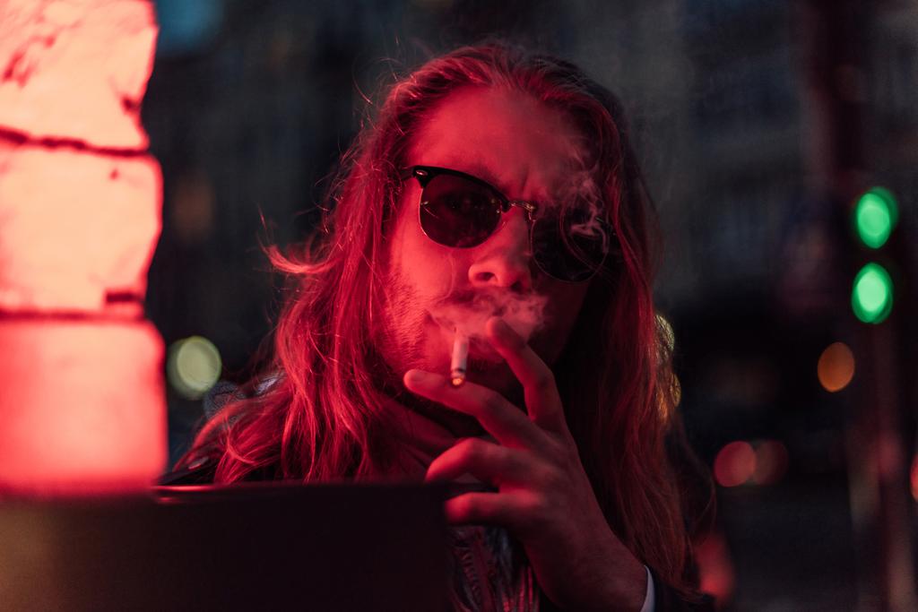 joven guapo en gafas de sol fumar cigarrillo bajo la luz roja en la calle
 - Foto, Imagen