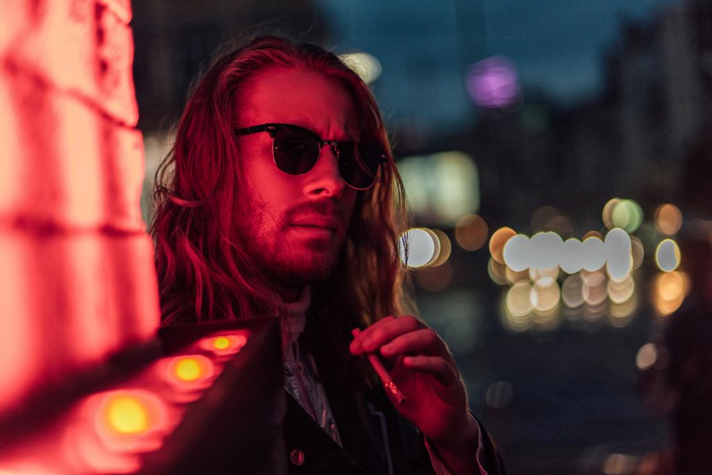 attraktiver junger Mann mit Sonnenbrille raucht Zigarette unter Rotlicht auf der Straße - Foto, Bild
