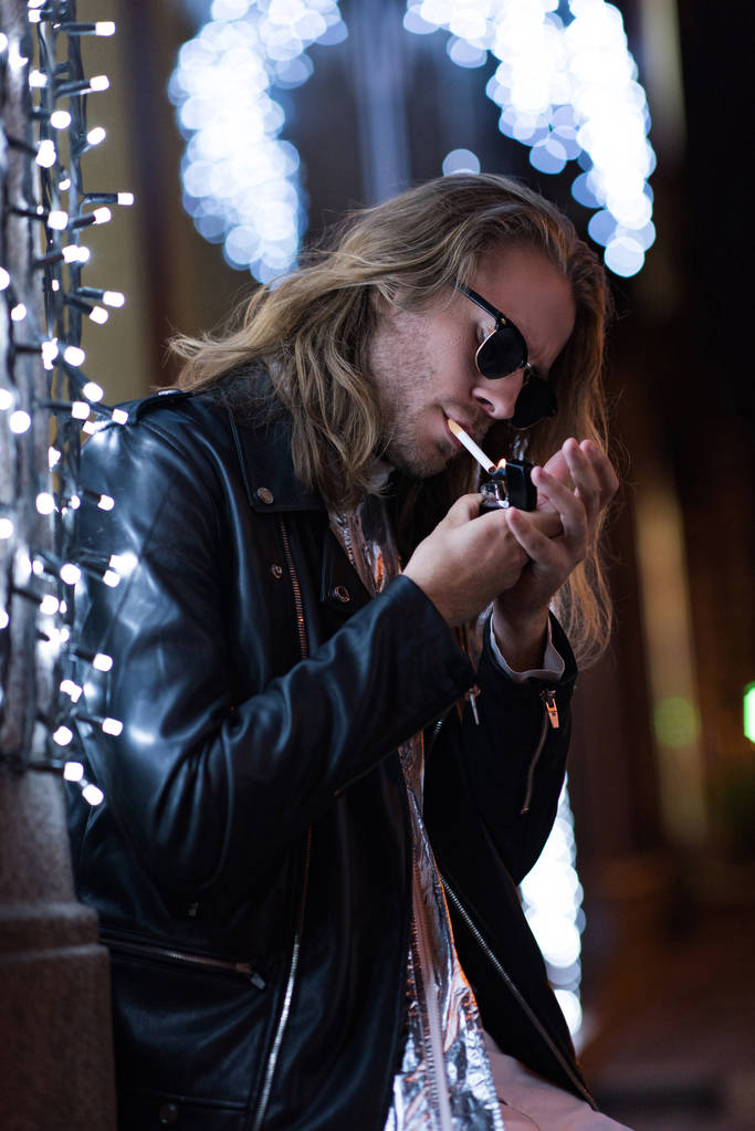 привабливий молодий чоловік в сонцезахисних окулярах і шкіряній куртці курить сигарету під гірляндою на міській вулиці вночі
 - Фото, зображення