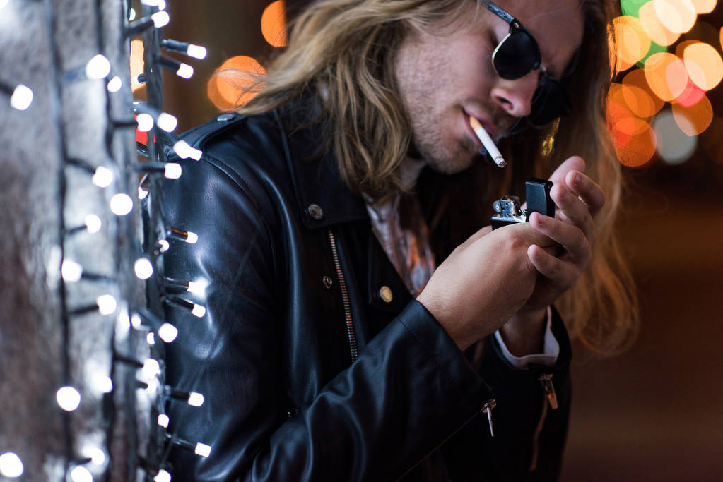 schöner junger Mann mit Sonnenbrille und Lederjacke, der nachts unter Girlanden auf der Straße Zigarette raucht - Foto, Bild