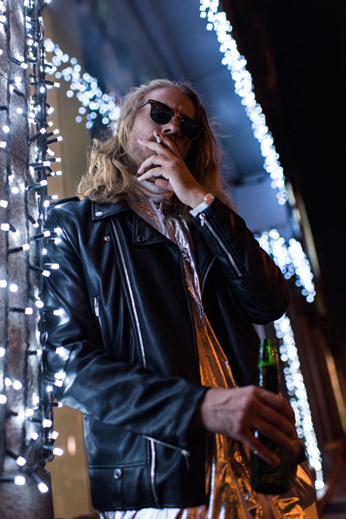 utcán éjszaka a csinos, fiatal férfi napszemüveg és bőr kabát üzem üveg sör és cigaretta a dohányzás alatt garland Alulnézet - Fotó, kép