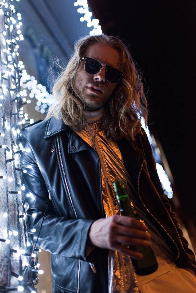 vista in basso di bel giovane uomo in occhiali da sole e giacca di pelle che tiene bottiglia di birra e in piedi sotto ghirlanda sulla strada di notte
 - Foto, immagini