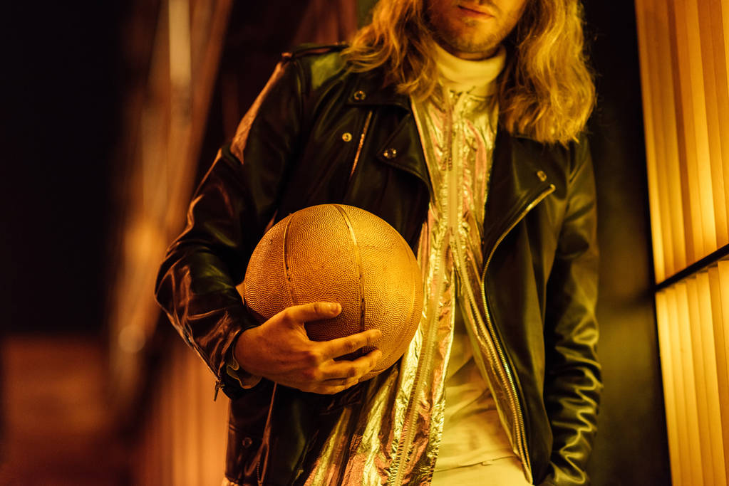 plan recadré de l'homme élégant en veste en cuir tenant ballon de basket-ball doré sous la lumière jaune dans la rue la nuit
 - Photo, image