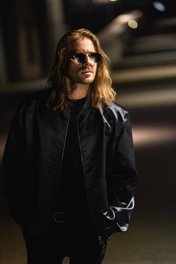 attractive young man in sunglasses and leather jacket on city street at night - Photo, Image
