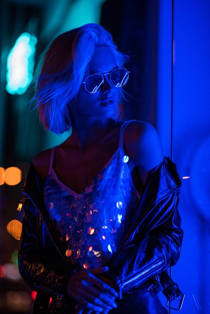 hermosa mujer joven en camiseta brillante y gafas de sol en la calle por la noche bajo la luz azul
 - Foto, Imagen