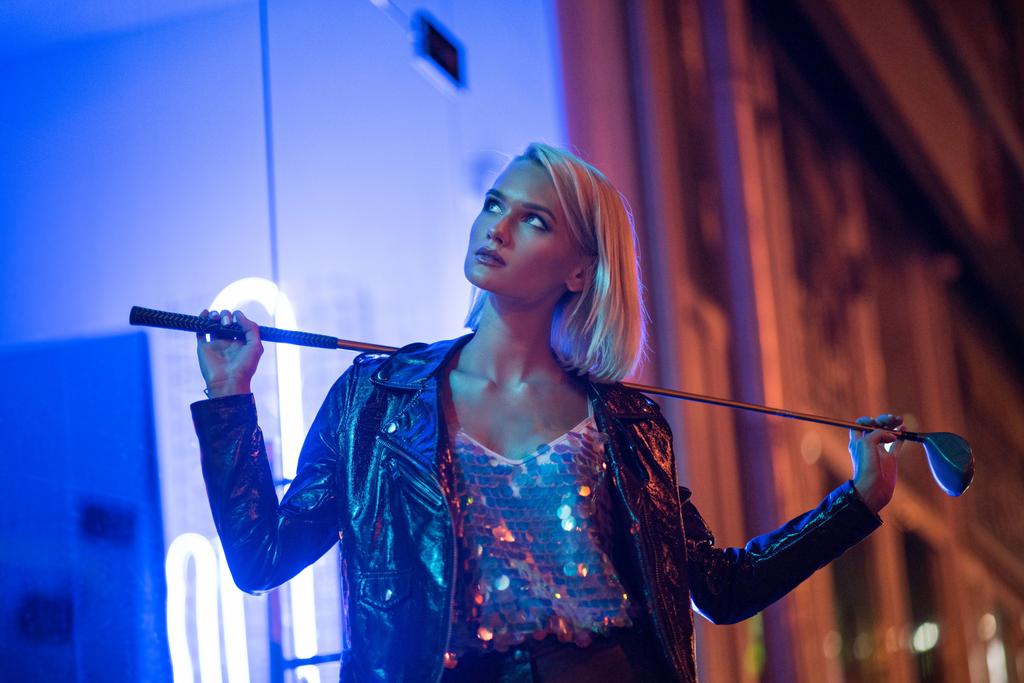 青いライトの下で夜に通りに立って押し続けるゴルフ クラブのレザー ジャケットの魅力的な若い女性 - 写真・画像