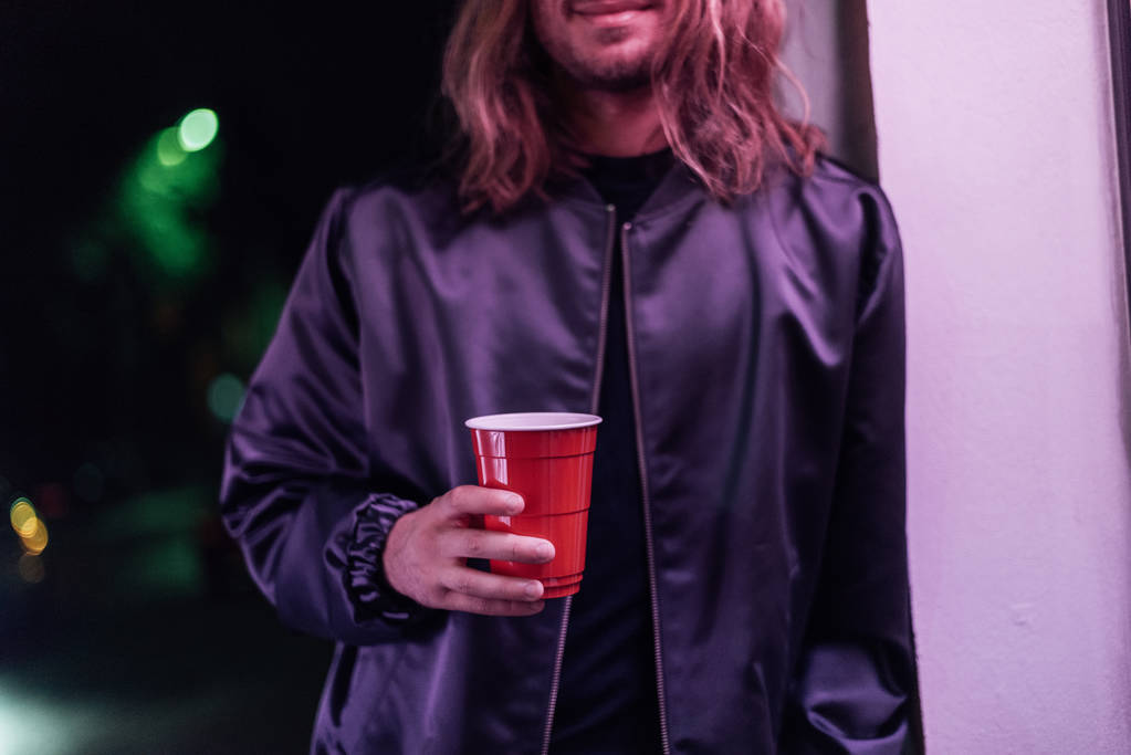 Genç adam kırpılmış kadeh içki sokakta gece pembe ışık altında kırmızı plastik bardak ile - Fotoğraf, Görsel