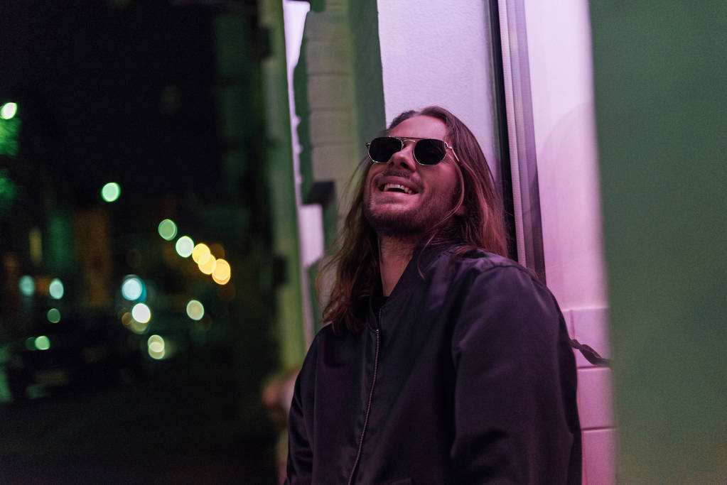 Smějící se mladý muž v kožené bundě a sluneční brýle na ulici v noci pod růžovým světlem - Fotografie, Obrázek