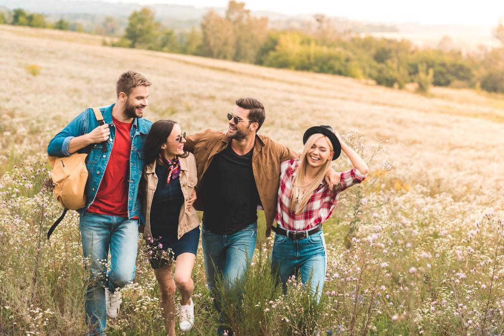 group of happy young friends embracing and walking by field together during trip - Photo, Image