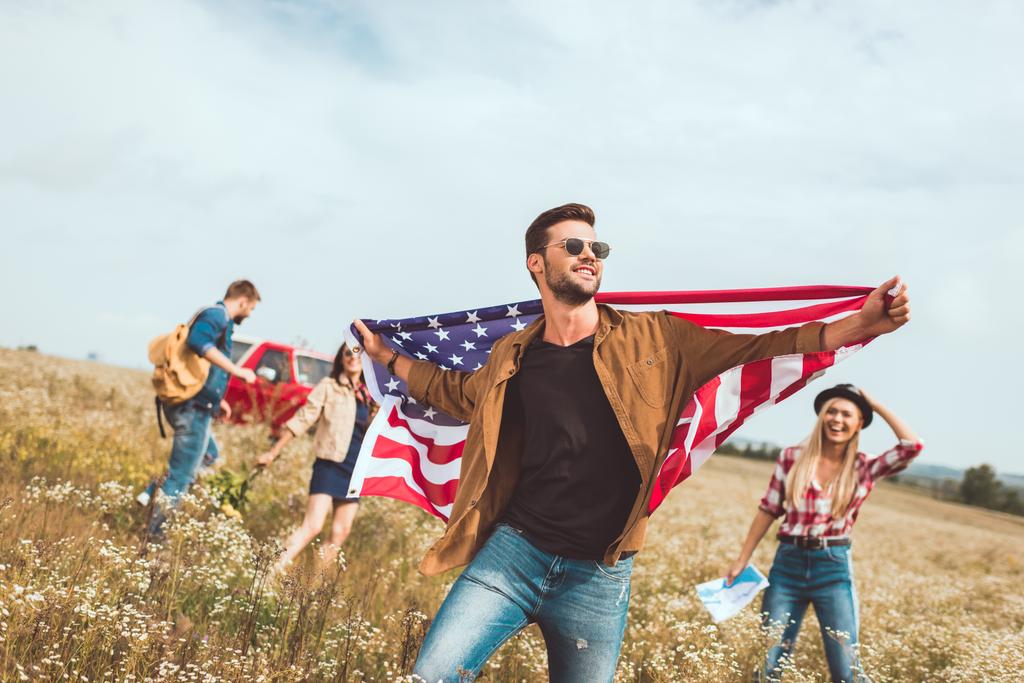όμορφος νεαρός άνδρας, κρατώντας την σημαία των ΗΠΑ και το περπάτημα από το πεδίο με τους φίλους κατά τη διάρκεια ταξιδιού - Φωτογραφία, εικόνα