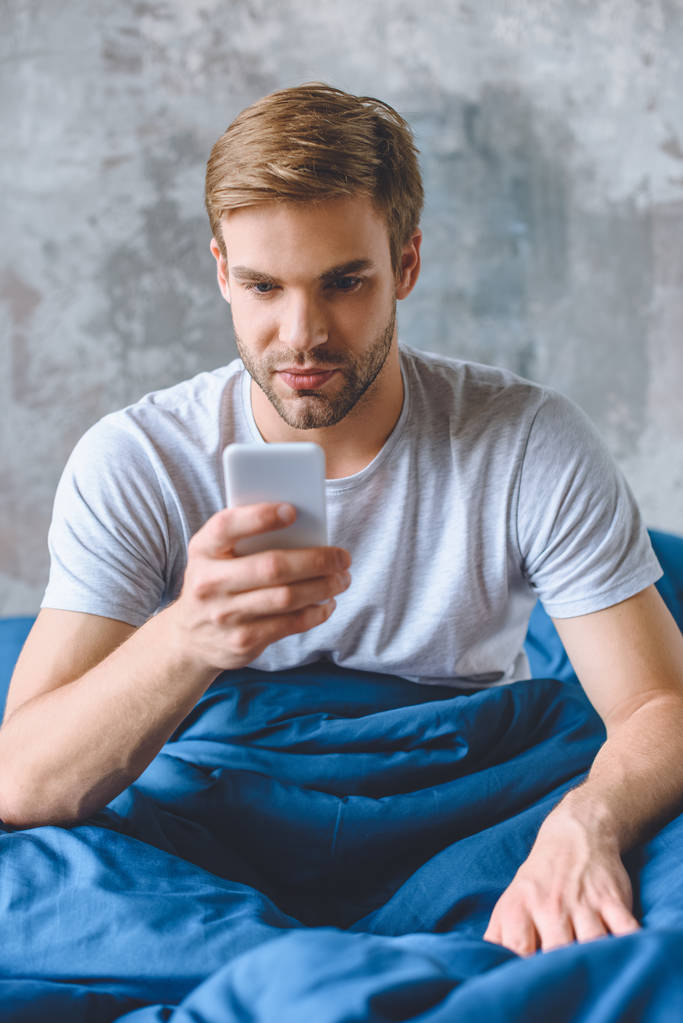 молодий чоловік використовує смартфон в ліжку вдома
 - Фото, зображення