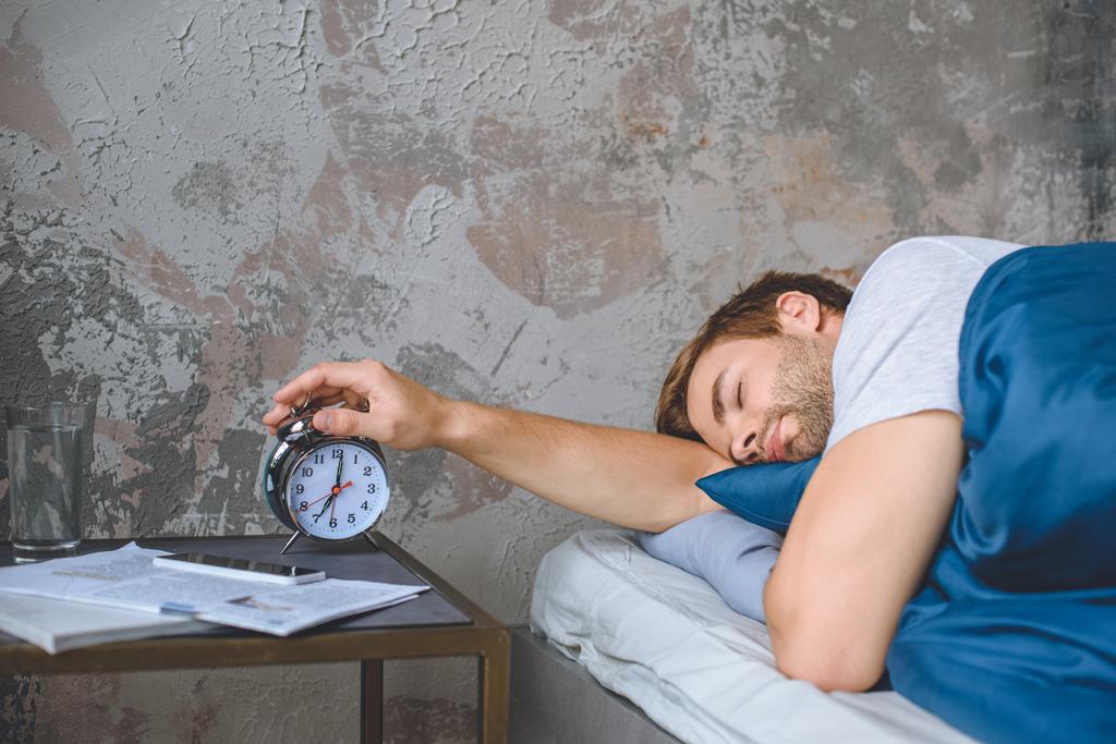 молодий сплячий чоловік в ліжку вимикає будильник вдома
  - Фото, зображення