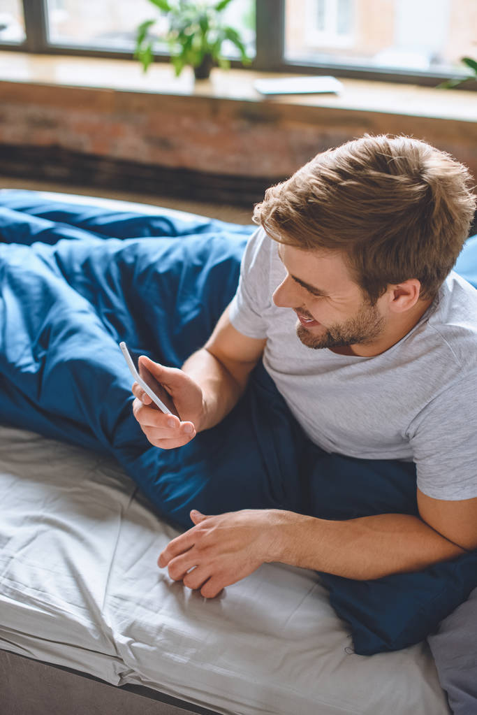 smartphone evde yatakta kontrol gülümseyen genç adamın yüksek açılı görünüş  - Fotoğraf, Görsel