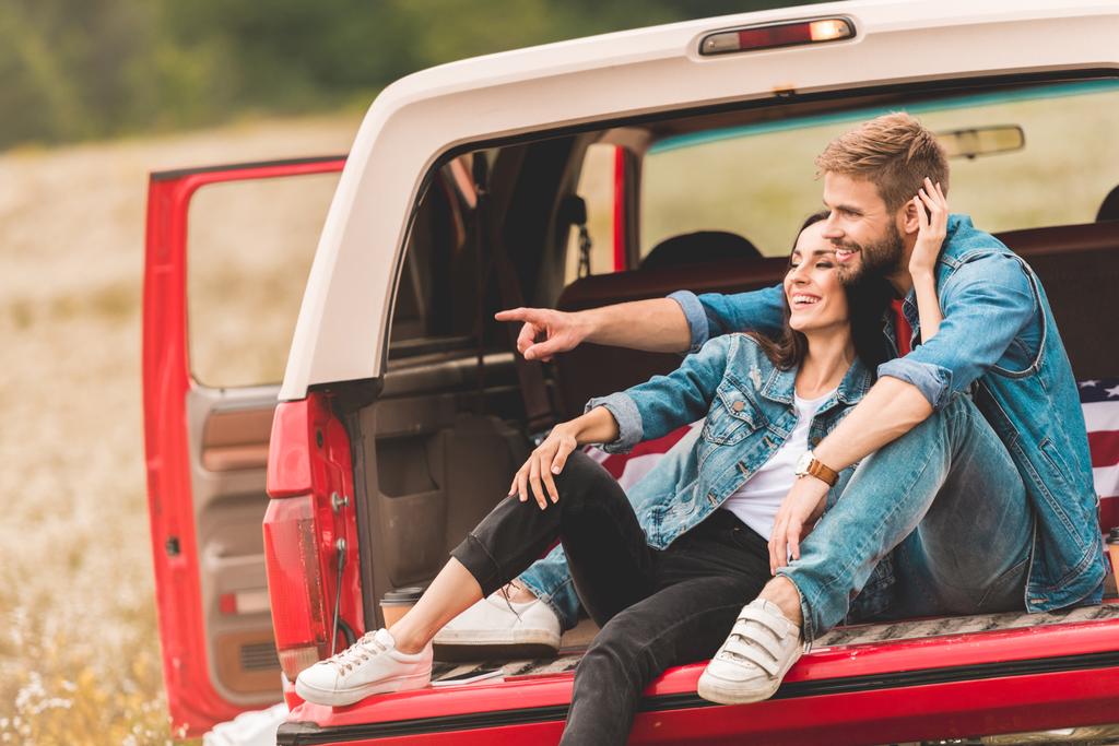 beautiful young couple relaxing in car trunk during trip and pointing somewhere - Photo, Image