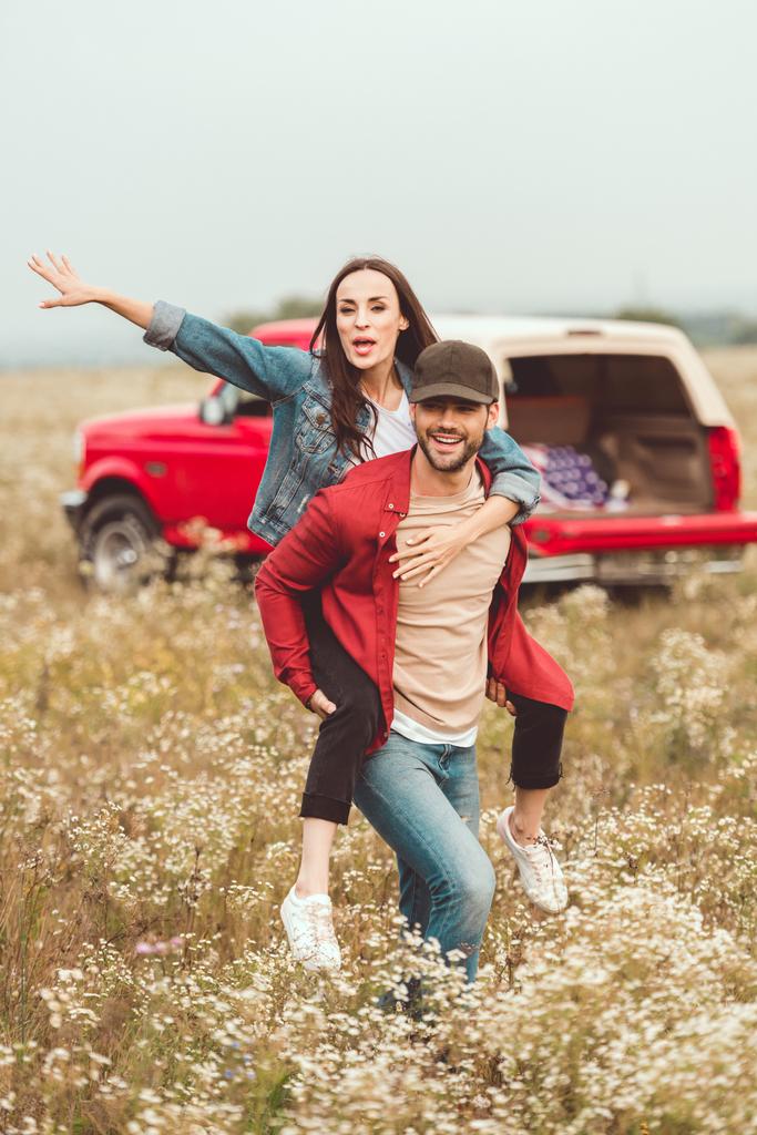 felice giovane donna a cavalluccio sul fidanzato in campo floreale con auto offuscata sullo sfondo
 - Foto, immagini