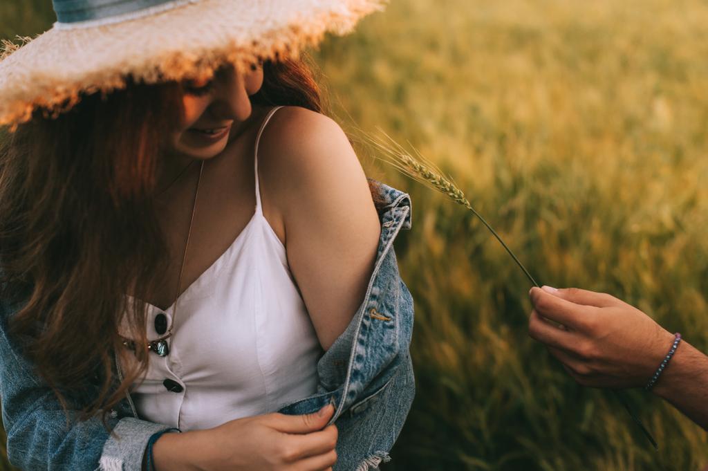 привлекательная улыбающаяся девушка в соломенной шляпе сидит на лугу, в то время как парень трогает ее пшеничным ухом к плечу
 - Фото, изображение