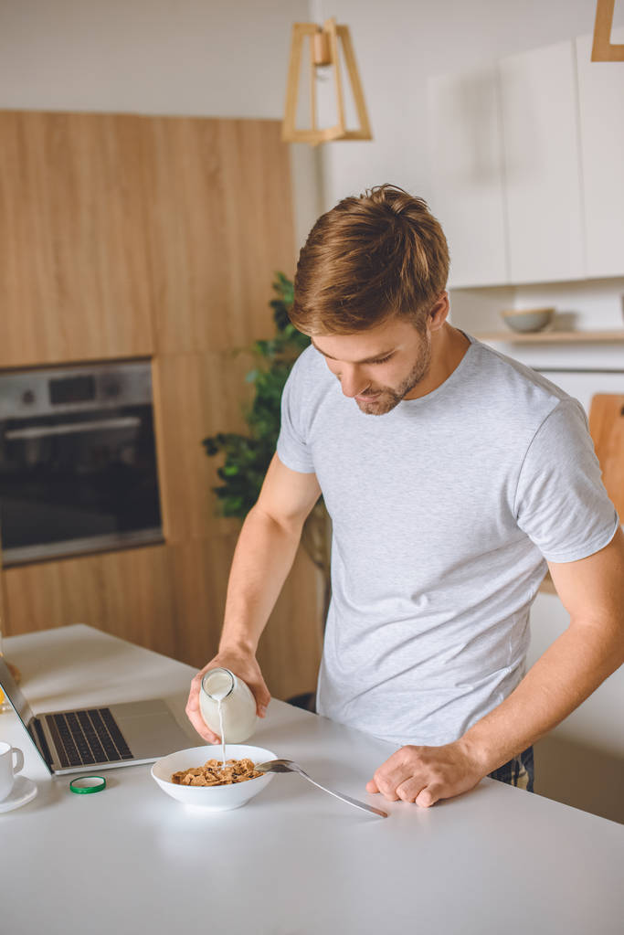 Junger Mann gießt am Küchentisch mit Laptop Milch in Teller mit Flocken  - Foto, Bild