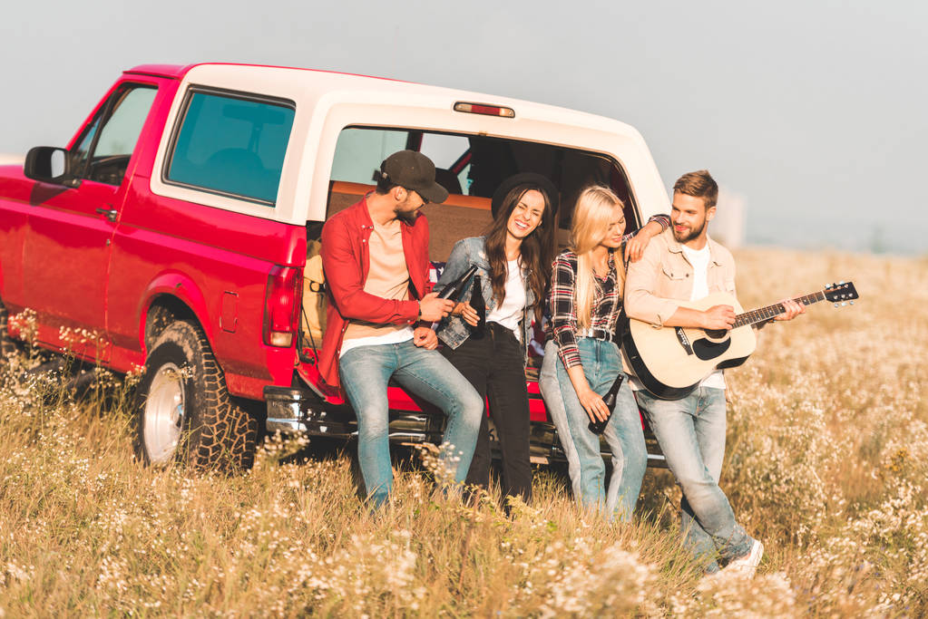 Gruppe fröhlicher junger Leute trinkt Bier und spielt Gitarre, während sie es sich im Kofferraum eines Autos auf einem Blumenfeld gemütlich macht - Foto, Bild
