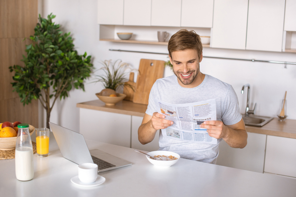 smiling young man reading newspaper at kitchen table with breakfast and laptop  - Photo, Image