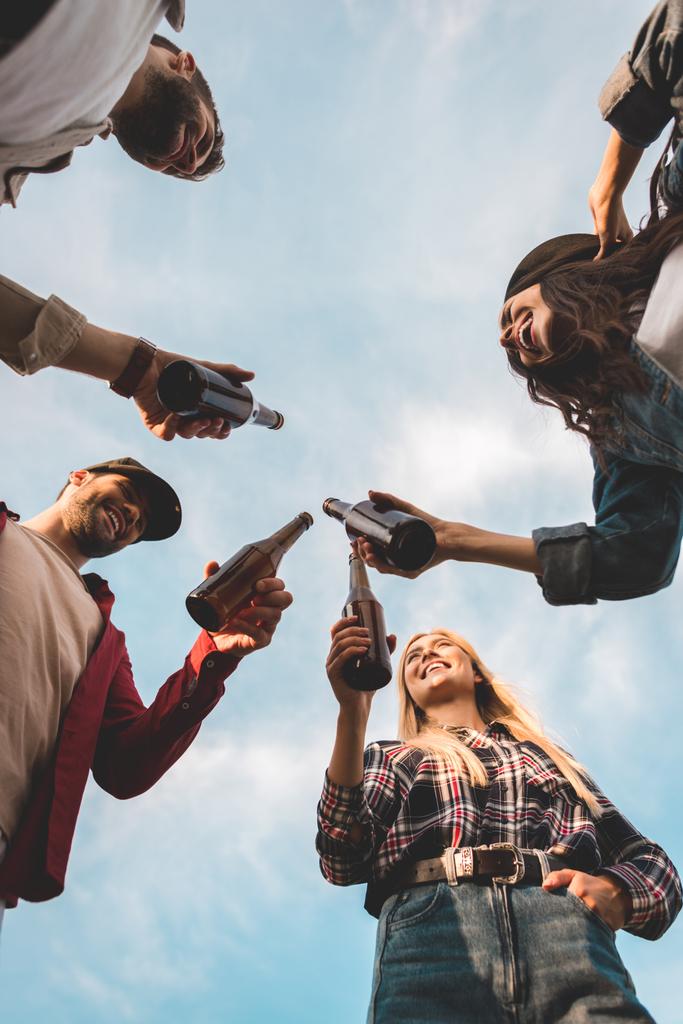 κάτω όψη ομάδας ευχαριστημένοι νέοι τσούγκριζαν μπουκάλια μπύρα μπροστά από το συννεφιασμένο ουρανό - Φωτογραφία, εικόνα