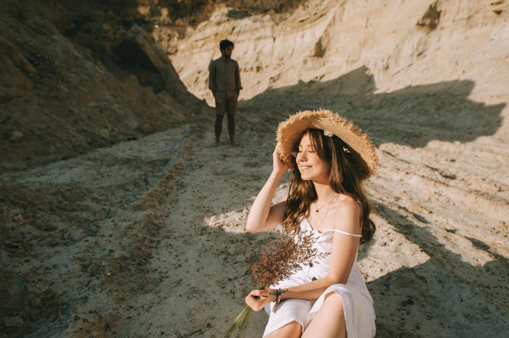 Gelukkig jonge vrouw in stro hoed met bloemen boeket zitten in zand canyon met vriendje achter - Foto, afbeelding