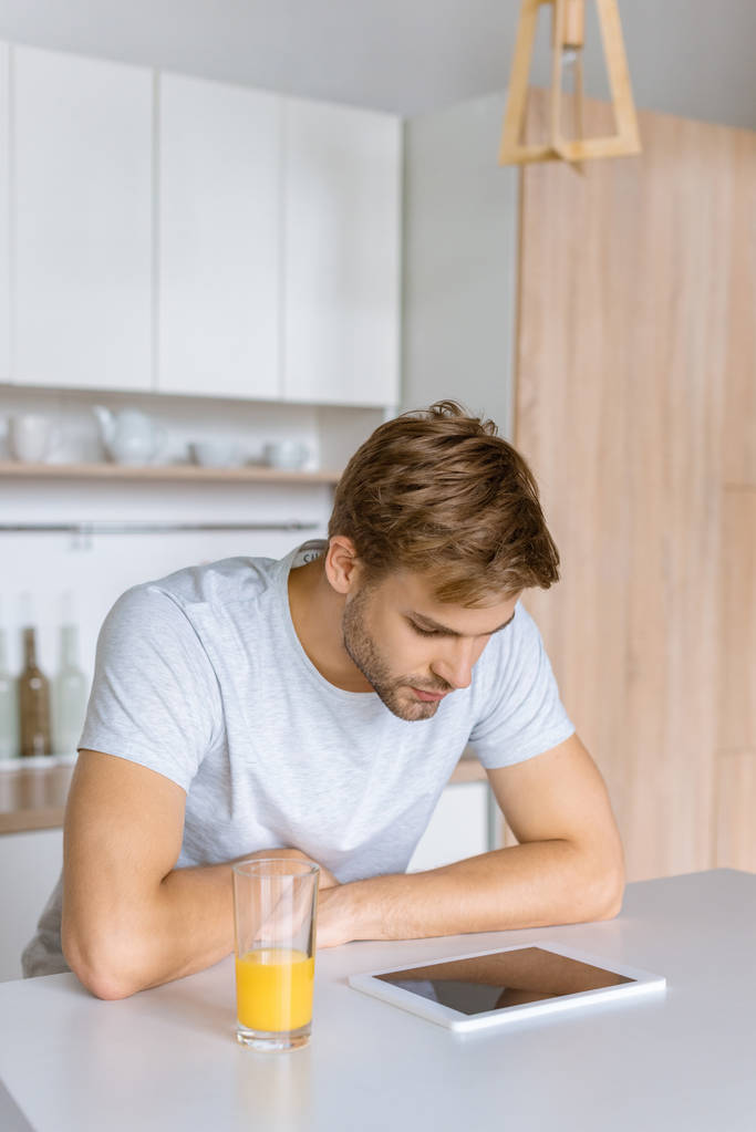 νεαρός άνδρας κοιτάζοντας ψηφιακή δισκίο στο τραπέζι της κουζίνας με φρέσκο χυμό  - Φωτογραφία, εικόνα