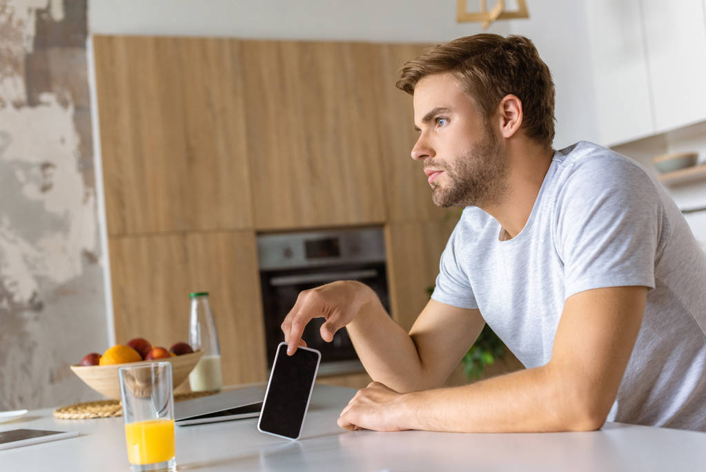 задумливий молодий чоловік зі смартфоном сидить за кухонним столом зі склянкою соку
  - Фото, зображення