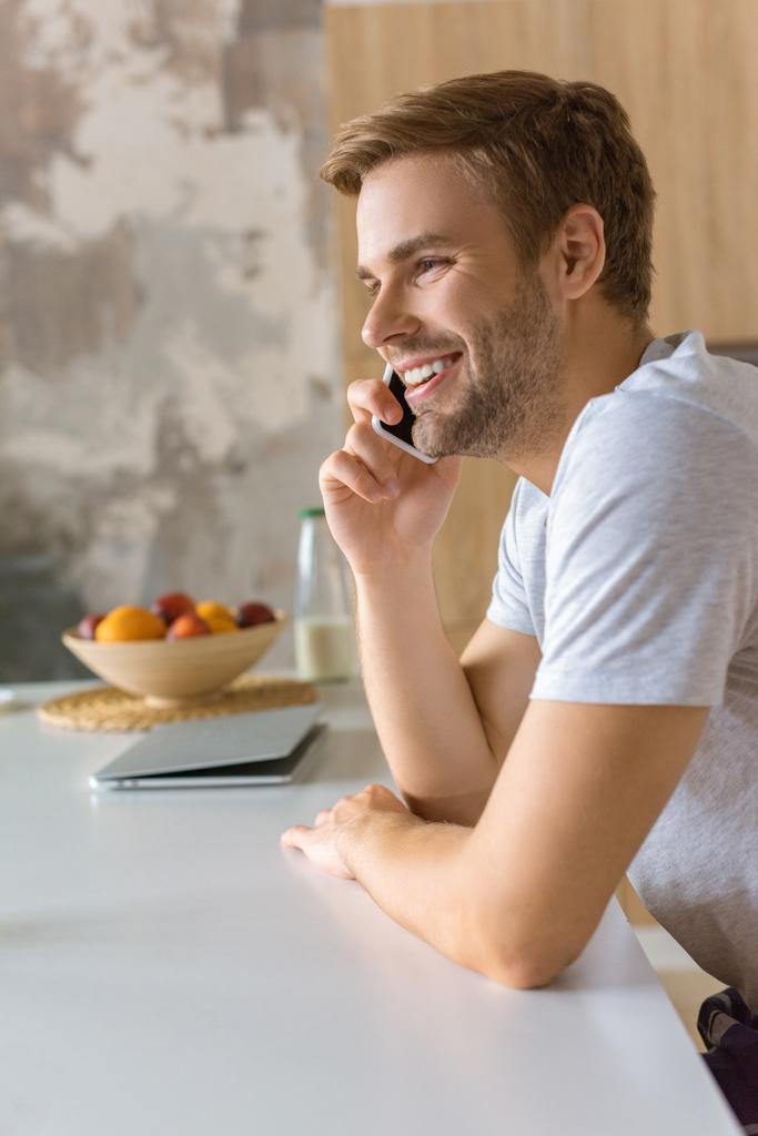selective focus of happy young man talking on smartphone at kitchen table  - Photo, Image