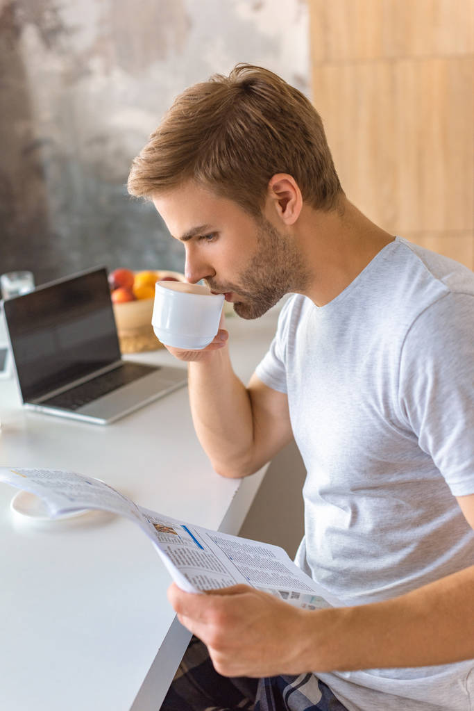 концентрированный молодой человек пьет кофе и читает газету на кухне
  - Фото, изображение