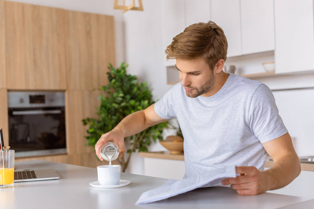 joven con el periódico vertiendo leche en la taza con café en la mesa de la cocina
   - Foto, Imagen