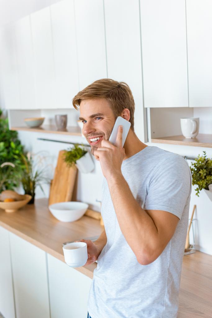 glimlachend jongeman met koffie op smartphone te praten tijdens de tijd van de ochtend op keuken  - Foto, afbeelding