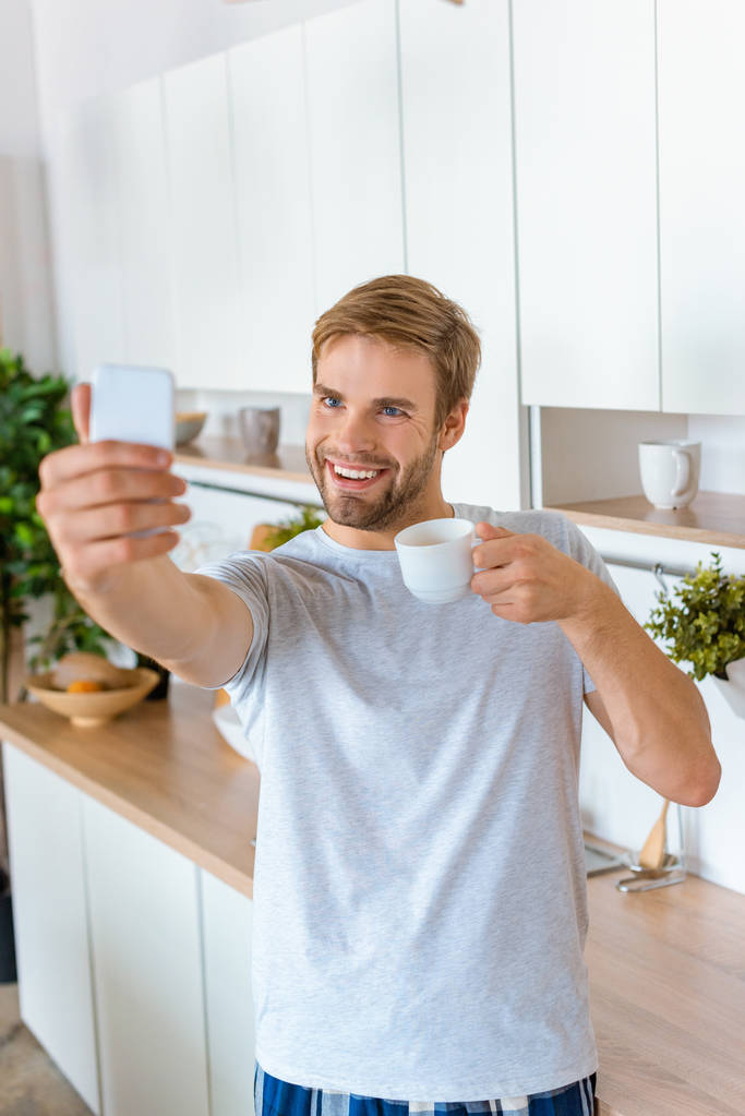 selfie キッチンでスマート フォンにコーヒー カップと笑顔の若い男 - 写真・画像