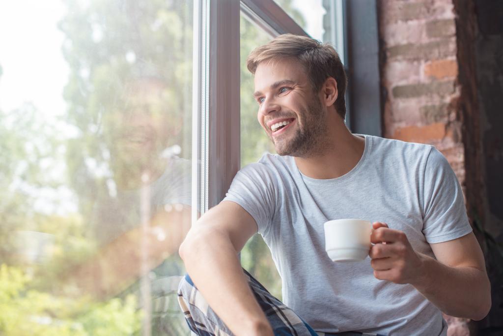 homme souriant regardant dans les fenêtres et assis avec une tasse de café sur le rebord de la fenêtre
  - Photo, image