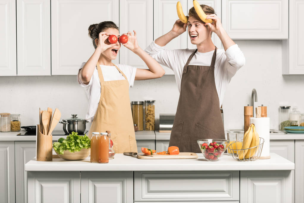 pareja joven divirtiéndose con plátanos y tomates en la cocina
 - Foto, Imagen