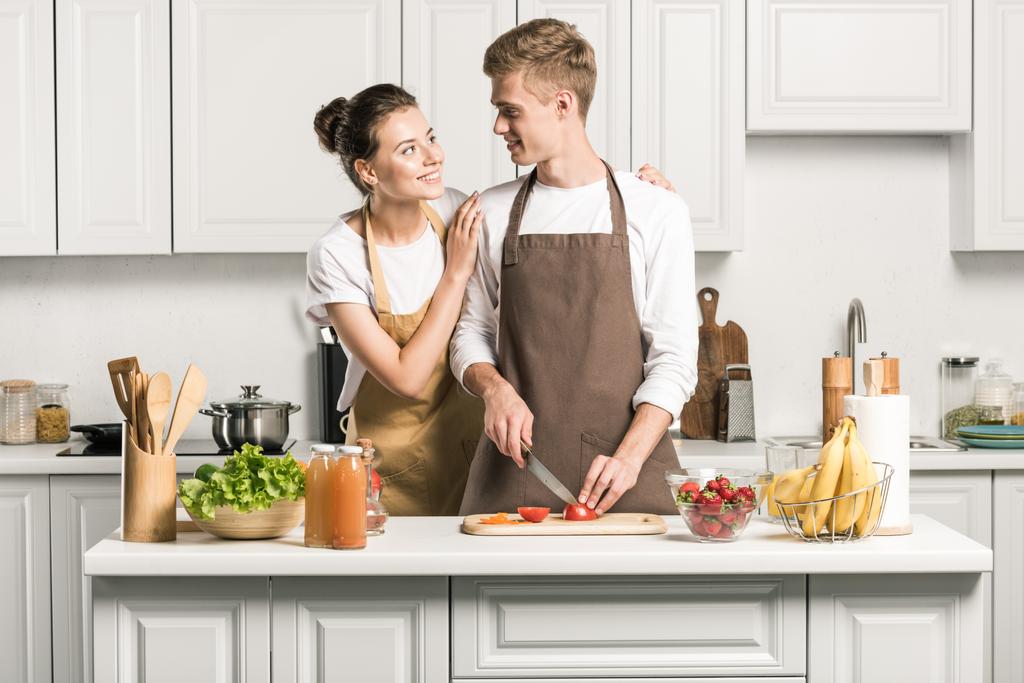 namorada abraçando namorado enquanto ele cortando tomates para salada na cozinha
 - Foto, Imagem