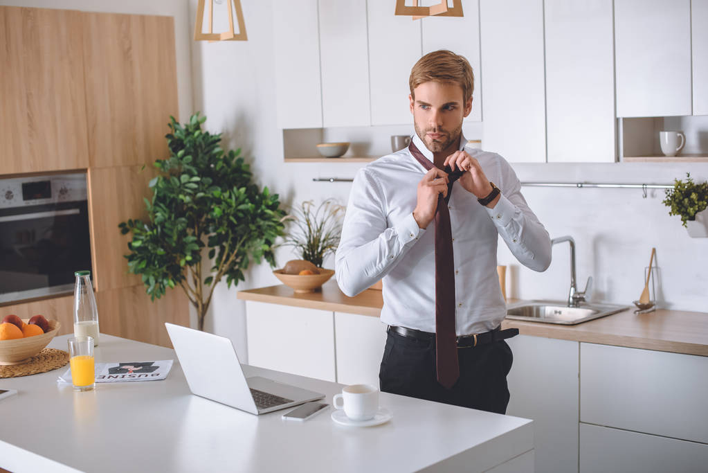 homme d'affaires en chemise blanche attachant cravate près de la table de cuisine avec ordinateur portable, smartphone et tasse à café
 - Photo, image