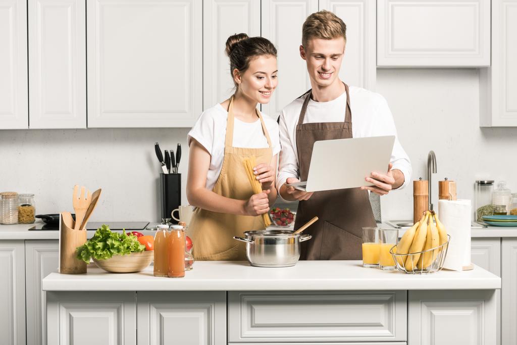 νεαρό ζευγάρι το μαγείρεμα των ζυμαρικών και χρήση φορητού υπολογιστή στην κουζίνα - Φωτογραφία, εικόνα