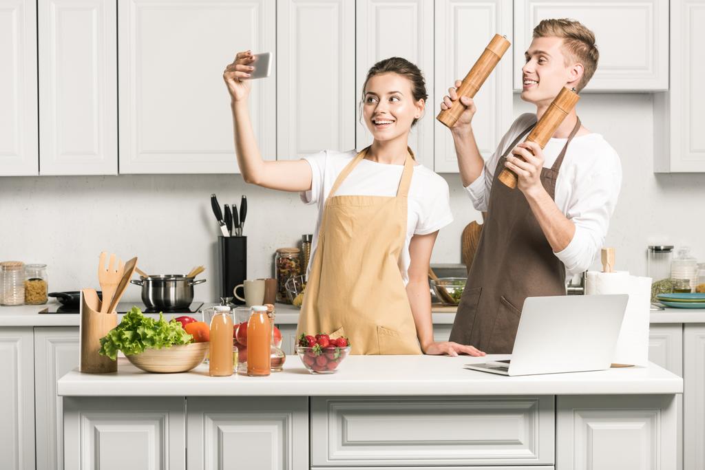 νεαρό ζευγάρι λαμβάνοντας αυτοπορτρέτα με smartphone κατά το μαγείρεμα στην κουζίνα - Φωτογραφία, εικόνα