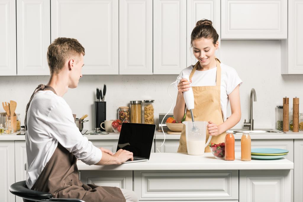 poikaystävä käyttää kannettavaa tietokonetta, tyttöystävä valmistelee pirtelöä keittiössä
 - Valokuva, kuva