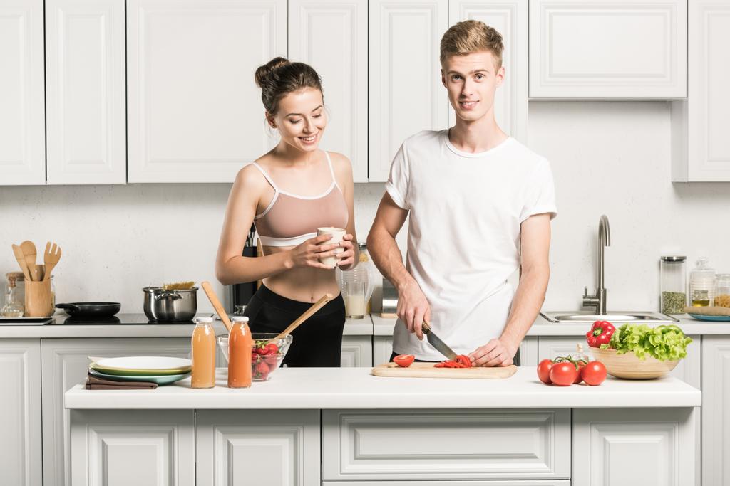 jovem casal cozinhar salada, namorado cortar tomates na cozinha
 - Foto, Imagem