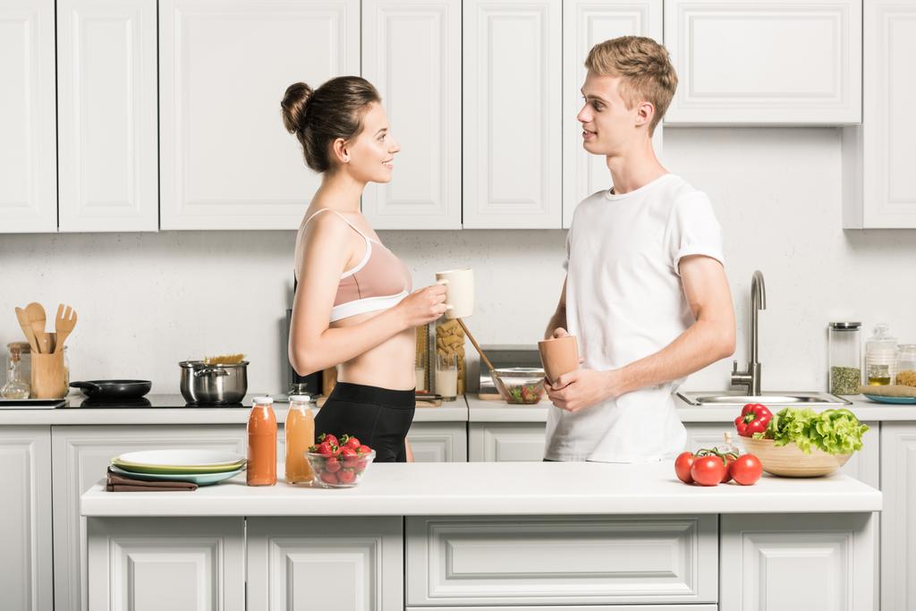 Seitenansicht eines jungen Paares, das Tassen hält und sich in der Küche anschaut - Foto, Bild