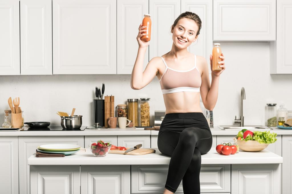 щаслива приваблива дівчина сидить на кухонній стійці і показує здоровий сік в пляшках
 - Фото, зображення