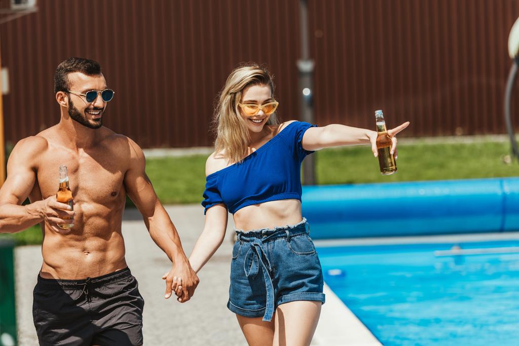 happy couple with bottles of beer holding hands, girl pointing at poolside - Photo, Image